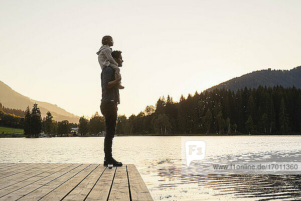 Vater nimmt seine kleine Tochter bei Sonnenuntergang am Ufer des Seestegs huckepack