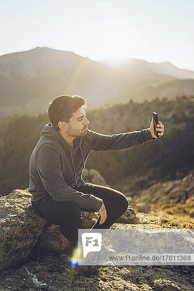 Junger Mann  der ein Selfie mit seinem Smartphone macht  während er an einem sonnigen Tag auf einem Berg sitzt