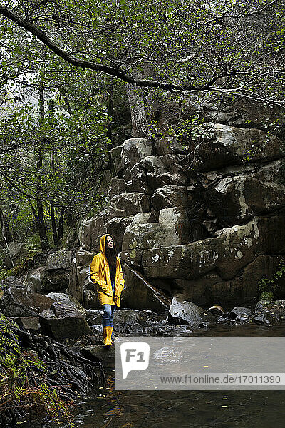Frau in gelbem Regenmantel mit geschlossenen Augen steht am Bach im Wald