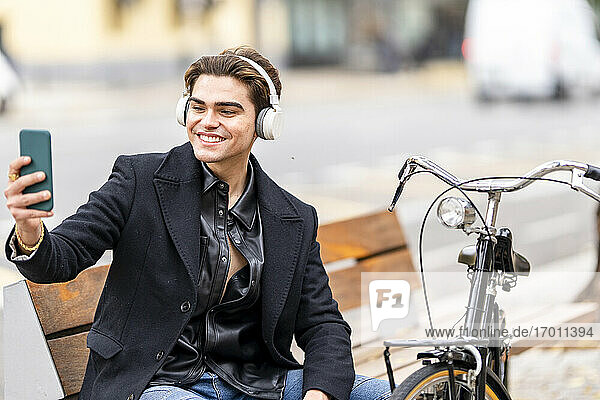 Lächelnder gut aussehender junger Mann  der ein Selfie mit seinem Smartphone macht  während er auf einem Fahrrad in der Stadt sitzt