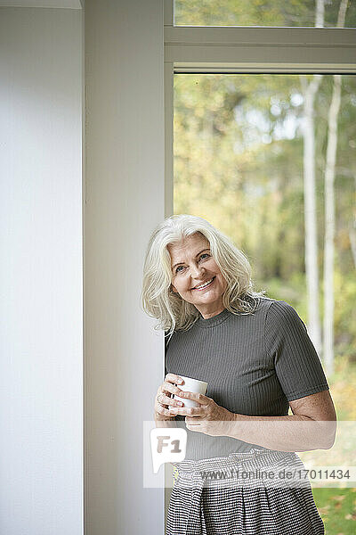 Lächelnde Frau im Ruhestand hält eine Tasse und lehnt sich an eine weiße Wand vor einem Fenster in einer Wohnung