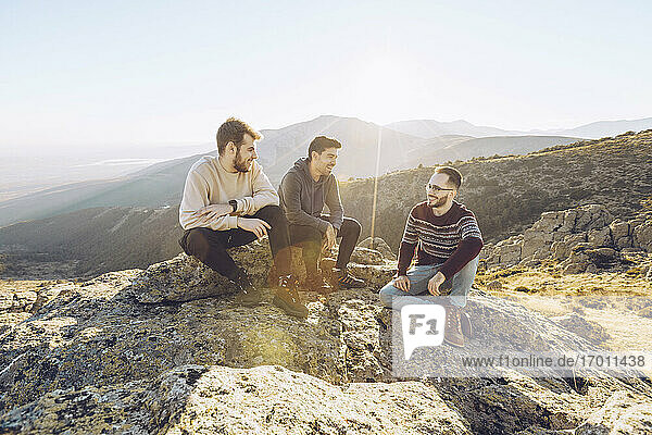 Male friends talking while sitting on mountain against clear sky during sunny day