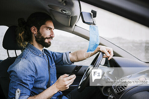 Lächelnder junger Mann sitzt im Auto und hält sein Smartphone während der COVID-19