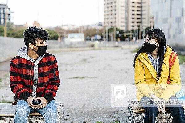 Paar mit Gesichtsschutzmaske sitzt in sozialer Distanz auf einer Bank während COVID-19