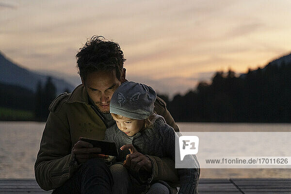 Vater und kleine Tochter spielen in der Abenddämmerung mit ihrem Smartphone auf einem Steg am Seeufer
