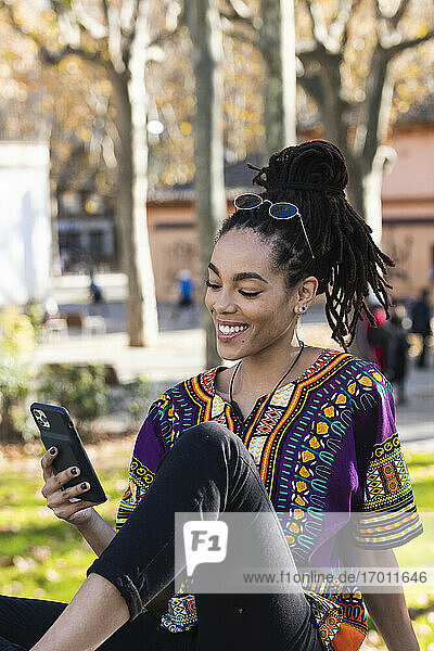 Lächelnde  modische junge Frau  die im Park sitzend ihr Smartphone benutzt