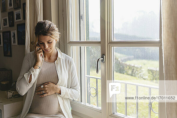 Junge schwangere Frau mit Hand auf dem Bauch  die zu Hause über ihr Smartphone spricht