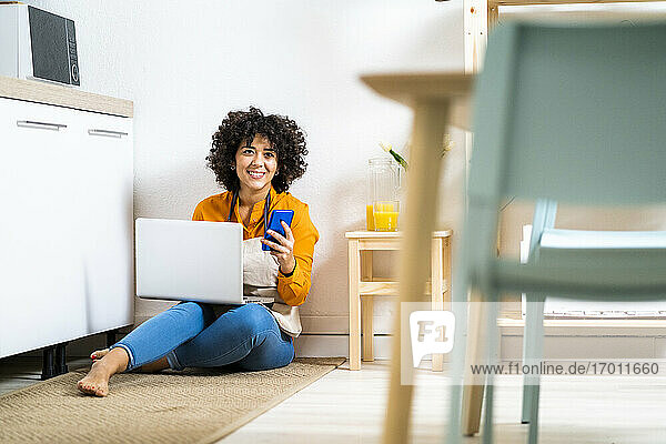 Smiling woman with laptop and mobile phone sitting on floor in kitchen at home