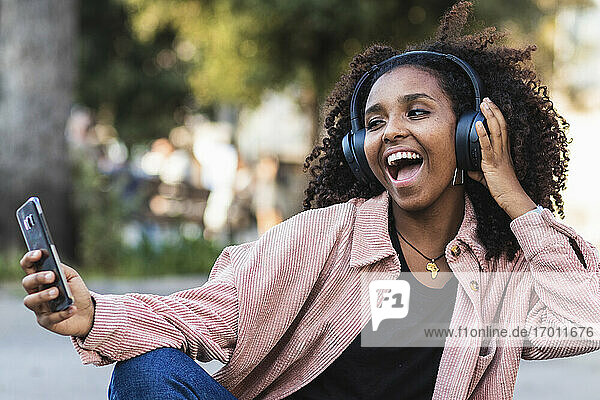Fröhliche junge Frau mit offenem Mund nimmt Selfie mit Kopfhörern in der Stadt