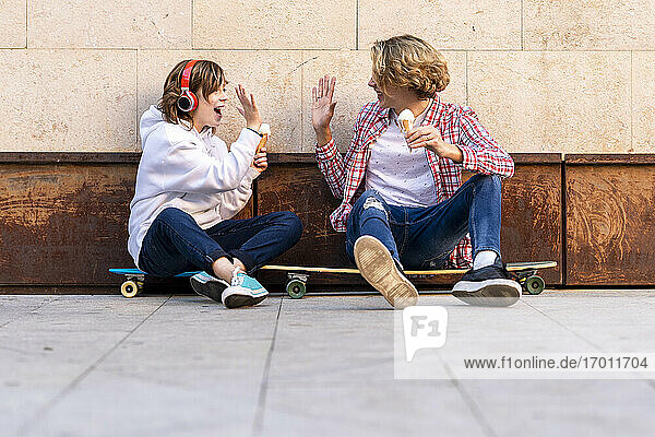 Lächelnder Junge und Mann mit Eiscreme geben sich High-Five  während sie auf einem Skateboard an der Wand sitzen