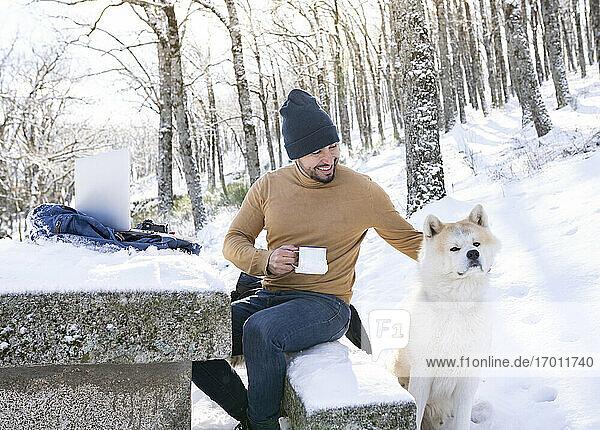 Lächelnder Mann hält Kaffeetasse und streichelt seinen Hund im verschneiten Wald
