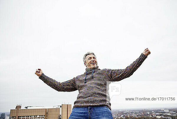 Happy man standing with arms outstretched against sky at rooftop