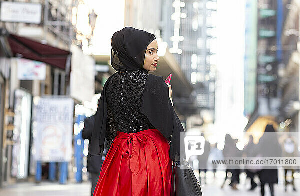 Porträt einer jungen schönen Frau mit schwarzem Hidschab  die auf dem Gehweg steht und über die Schulter in die Kamera schaut