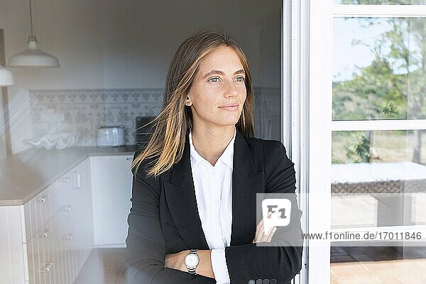 Thoughtful young businesswoman with arms crossed leaning on door while looking away