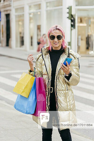 Lächelnde Frau  die Einkaufstüten trägt  während sie ein Mobiltelefon benutzt  in der Stadt stehend