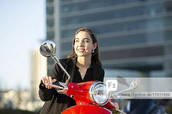 Lächelnde Geschäftsfrau schaut weg  während sie bei Sonnenuntergang auf einem Motorroller sitzt