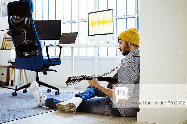 Male guitarist wearing knit hat playing guitar while sitting on floor at studio
