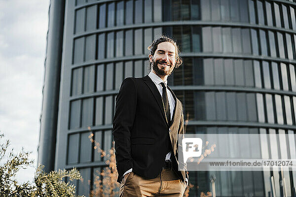 Young businessman with hands in pockets smiling while standing against building