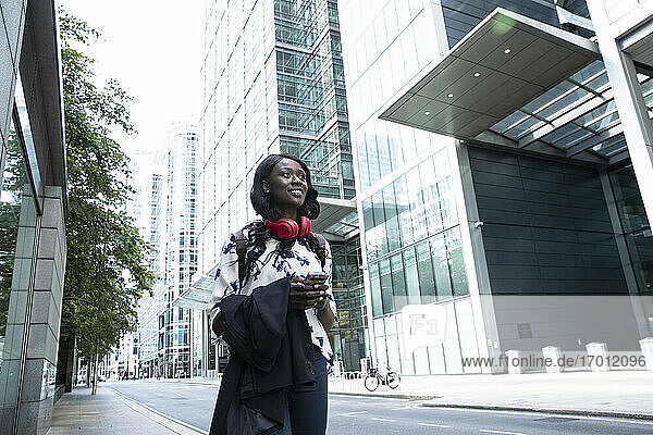 Junge Geschäftsfrau mit Kopfhörern und Mobiltelefon  die beim Spaziergang in der Stadt wegschaut