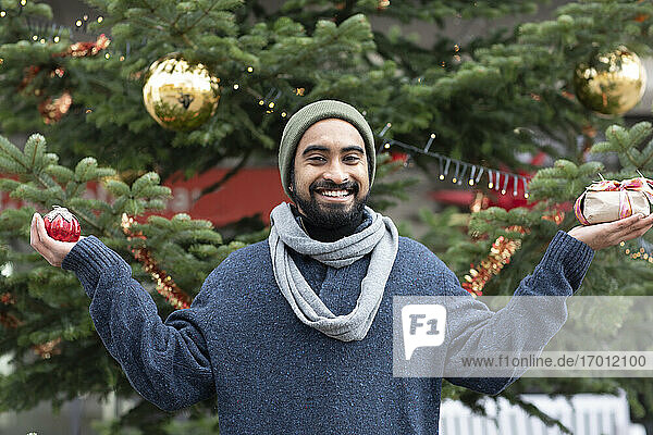 Lächelnder junger Mann  der ein Geschenk und ein Ornament vor einem Weihnachtsbaum hält