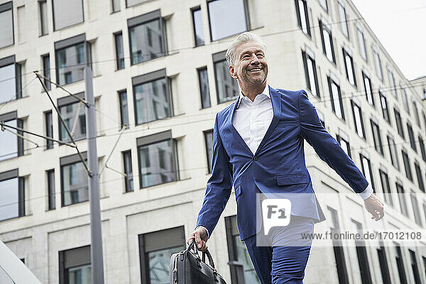 Lächelnder älterer männlicher Unternehmer mit Tasche  der gegen ein Gebäude in der Stadt läuft