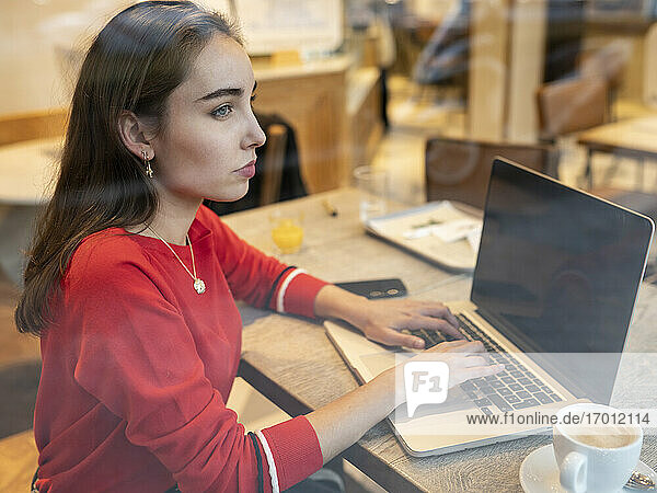Nachdenkliche Frau mit Laptop durch das Glas eines Cafés gesehen