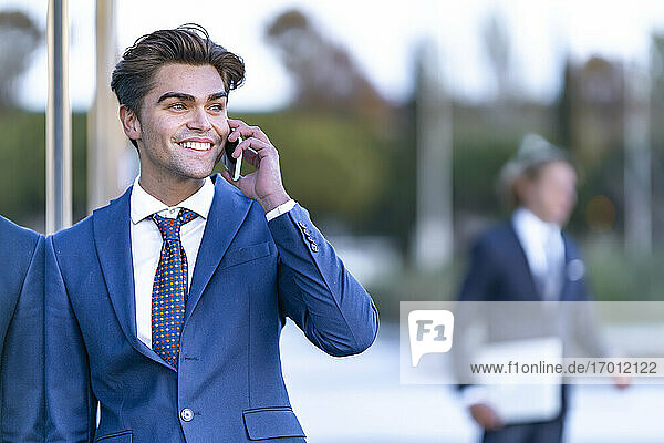 Happy businessman in suit leaning on glass while talking on smart phone