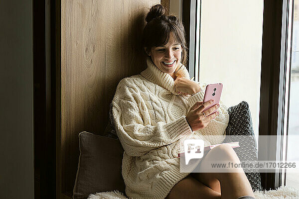 Woman in sweater smiling while video calling through mobile phone by window at home