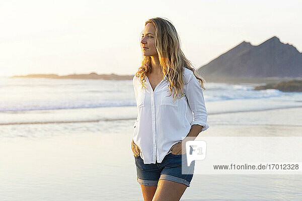 Beautiful woman with hands in pockets looking away while walking at beach