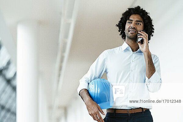 Junger männlicher Bauunternehmer mit Schutzhelm  der im Flur eines Büros mit einem Mobiltelefon spricht