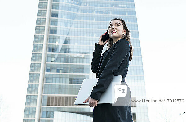 Young businesswoman with laptop talking on mobile phone while standing in city
