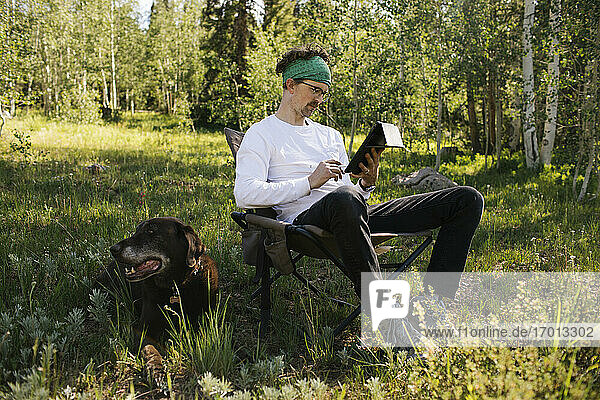 USA  Utah  Uinta National Park  Mann mit Hund sitzt auf einer Wiese im Wald  benutzt Tablet