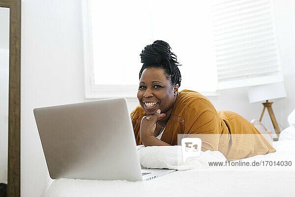 Porträt einer lächelnden Frau auf dem Bett liegend vor einem Laptop