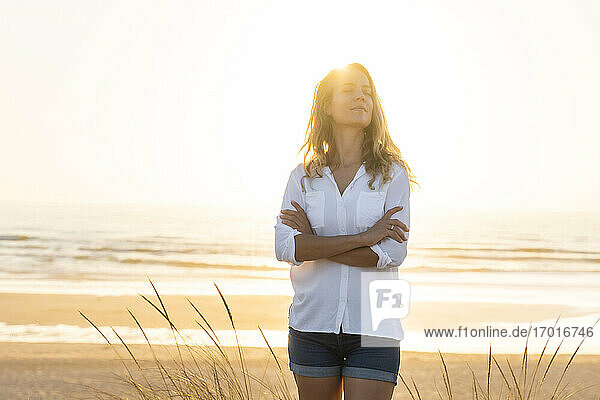 Mid adult woman standing wit arms crossed at beach during sunset