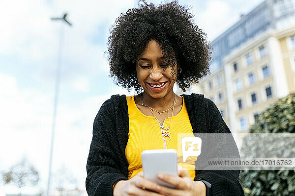 Lächelnde schöne Frau  die ein Mobiltelefon benutzt  während sie gegen den Himmel sitzt