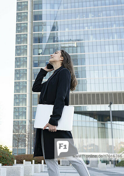 Businesswoman with laptop smiling while talking on mobile phone walking in city