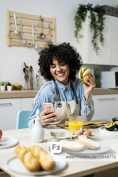 Porträt einer jungen Frau  die am Küchentisch mit einem Sandwich in der Hand ein Smartphone benutzt