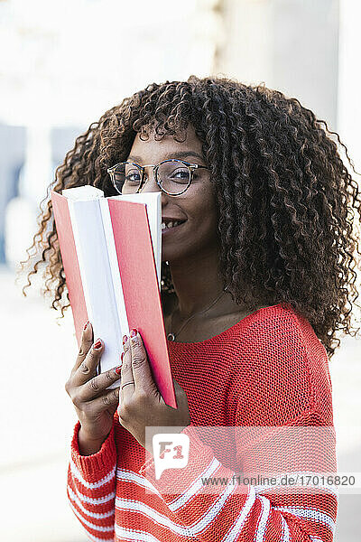 Happy young woman in warm clothes covering face with book
