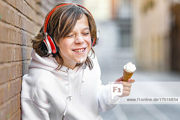 Lächelnder Junge mit Kopfhörern  der ein Eis in der Hand hält und sich an die Wand lehnt
