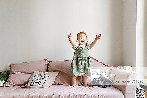 Lächelndes kleines Mädchen  das mit erhobenen Armen auf dem Sofa steht