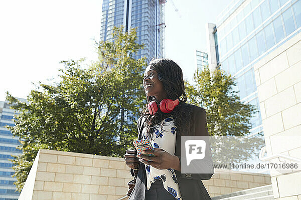 Lächelnde Geschäftsfrau mit Kopfhörern und Rucksack  die ein Mobiltelefon benutzt  während sie in der Stadt steht