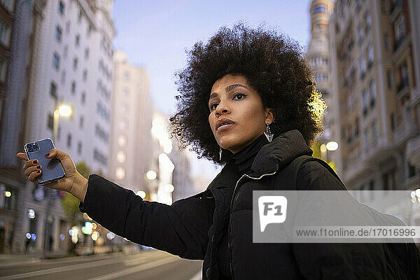 Afro-Frau mit Handy in der Hand  während sie nachts auf einer Straße in der Stadt eine Mitfahrgelegenheit sucht
