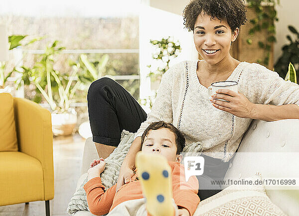 Frau mit Kaffeetasse  die lächelnd neben einem Jungen sitzt  der sich zu Hause auf dem Sofa ausruht