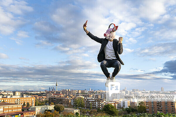 Male entrepreneur wearing pig mask taking selfie from smart phone while jumping in city against sky