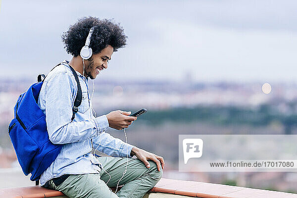 Smiling young man listening music and using smart phone on terrace against sky