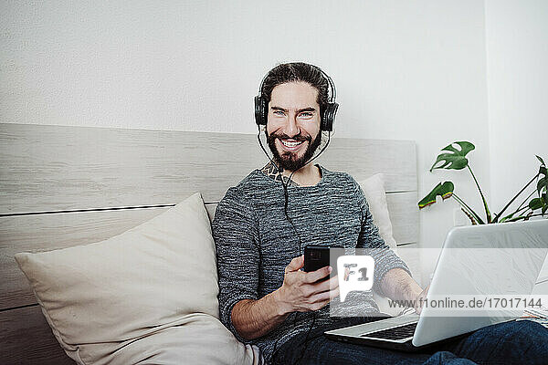 Lächelnder Mann mit Kopfhörern  der ein Mobiltelefon benutzt  während er mit einem Laptop auf dem Bett zu Hause sitzt