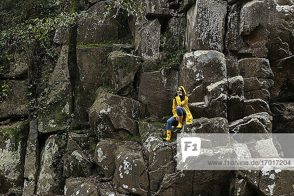 Nachdenkliche junge Frau in gelbem Regenmantel sitzt auf einem Felsen im Wald