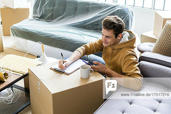 Lächelnder Mann mit Mobiltelefon  der über einem Karton in seinem neuen Zuhause auf Papier schreibt