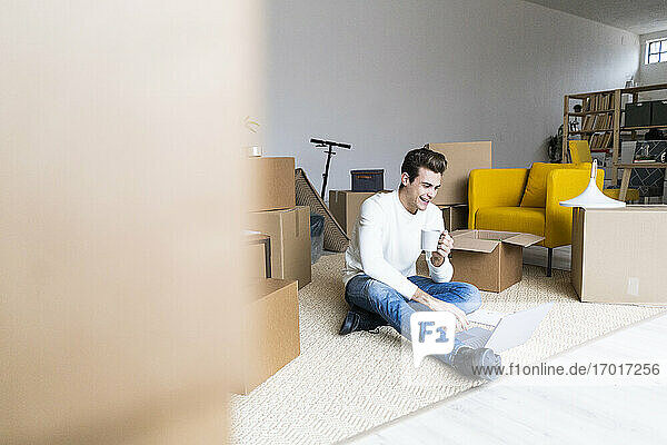 Smiling young man having coffee while using laptop in new loft apartment