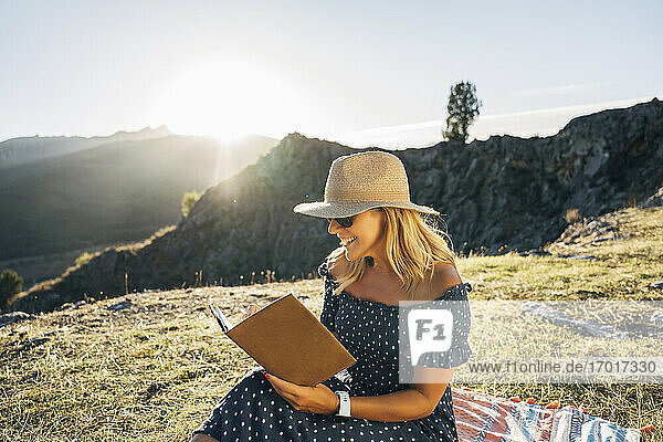 Glückliche Frau  die ein Buch liest  während sie auf einer Picknickdecke vor den Bergen an einem sonnigen Tag sitzt